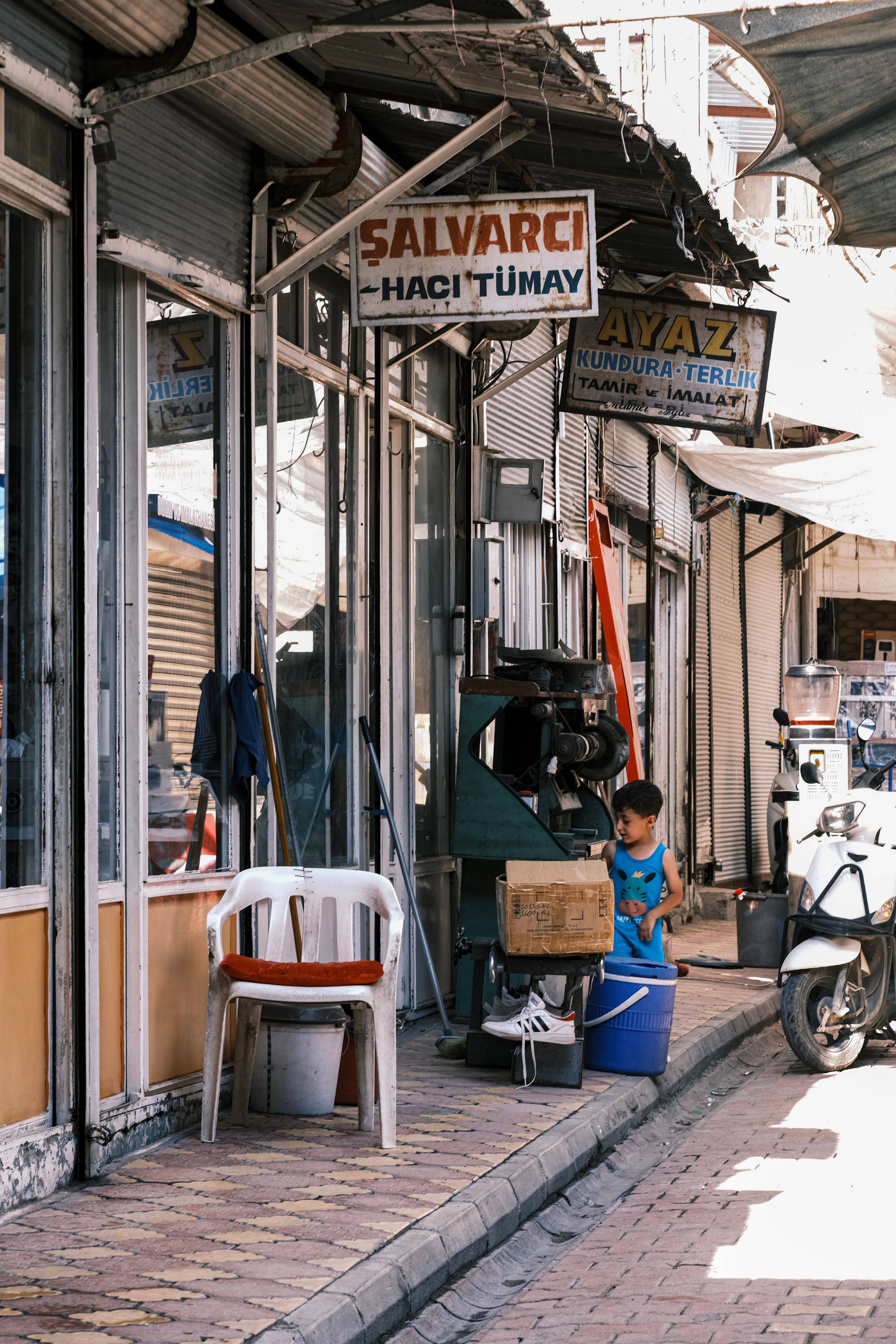 A child stands near shops on a narrow street in a Turkish market, surrounded by signs and scooters.