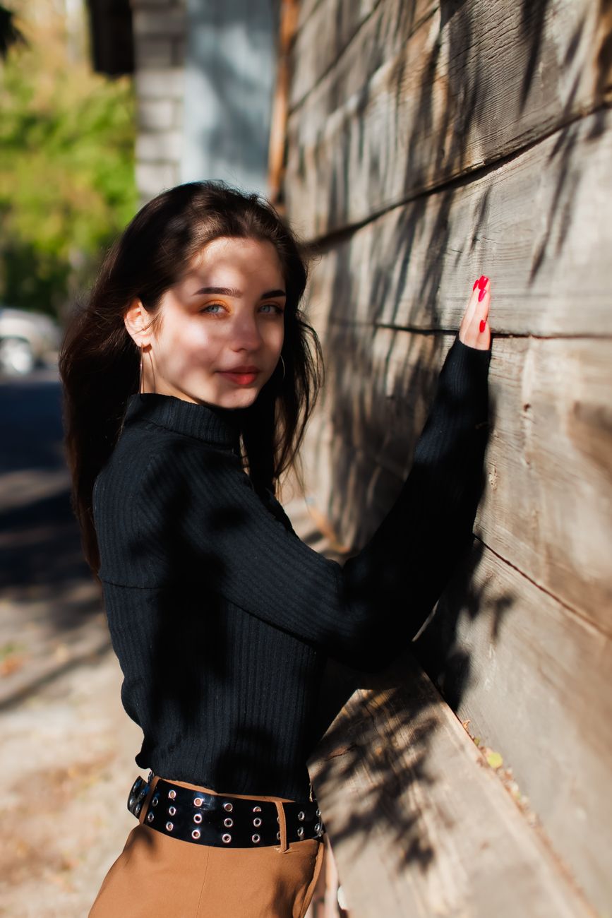 Woman in a black sweater poses outdoors against a rustic wooden wall on a sunny day.