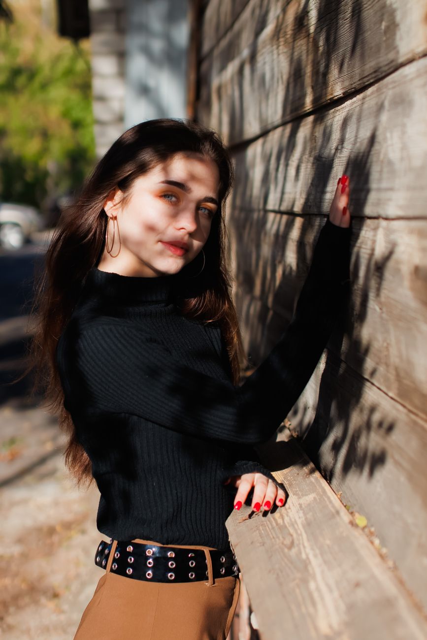 A young woman in a black top leans against a rustic wooden wall in sunlight, casting shadows.