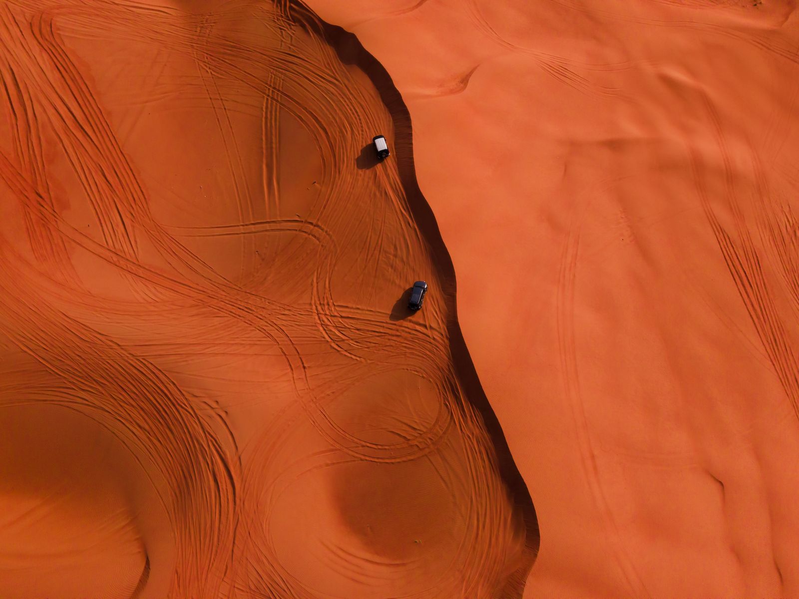 Aerial view of two small 4x4 vehicles crossing vast treeless orange sand dunes with no natural winch anchor points anywhere