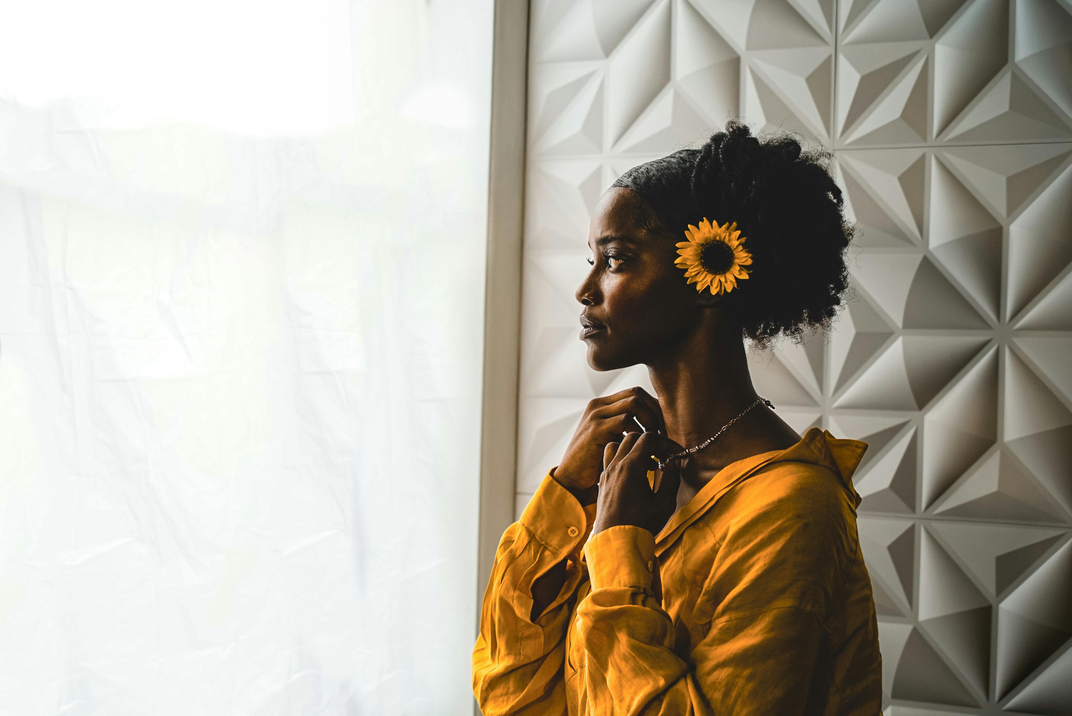 Elegant portrait of a woman by a window, sunflower in hair, thoughtful gaze.