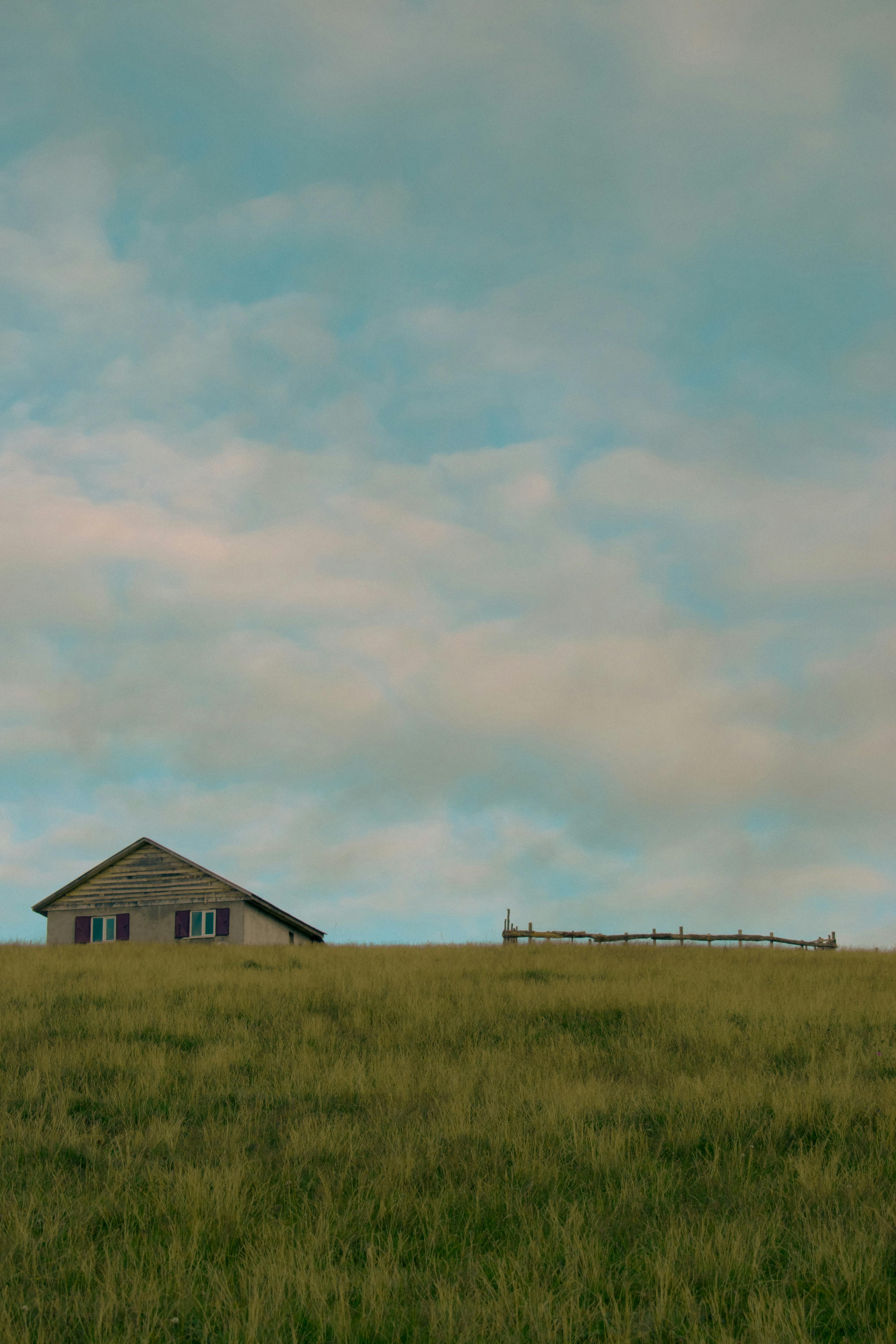 A rustic wooden cabin sits isolated on a vast green meadow under a blue sky with fluffy clouds, evoking serenity.