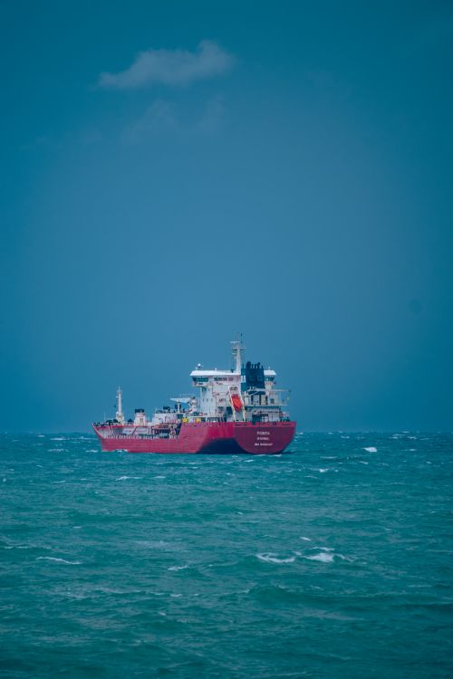Free A solitary red cargo ship sails on the turquoise sea under a clear blue sky. Stock Photo