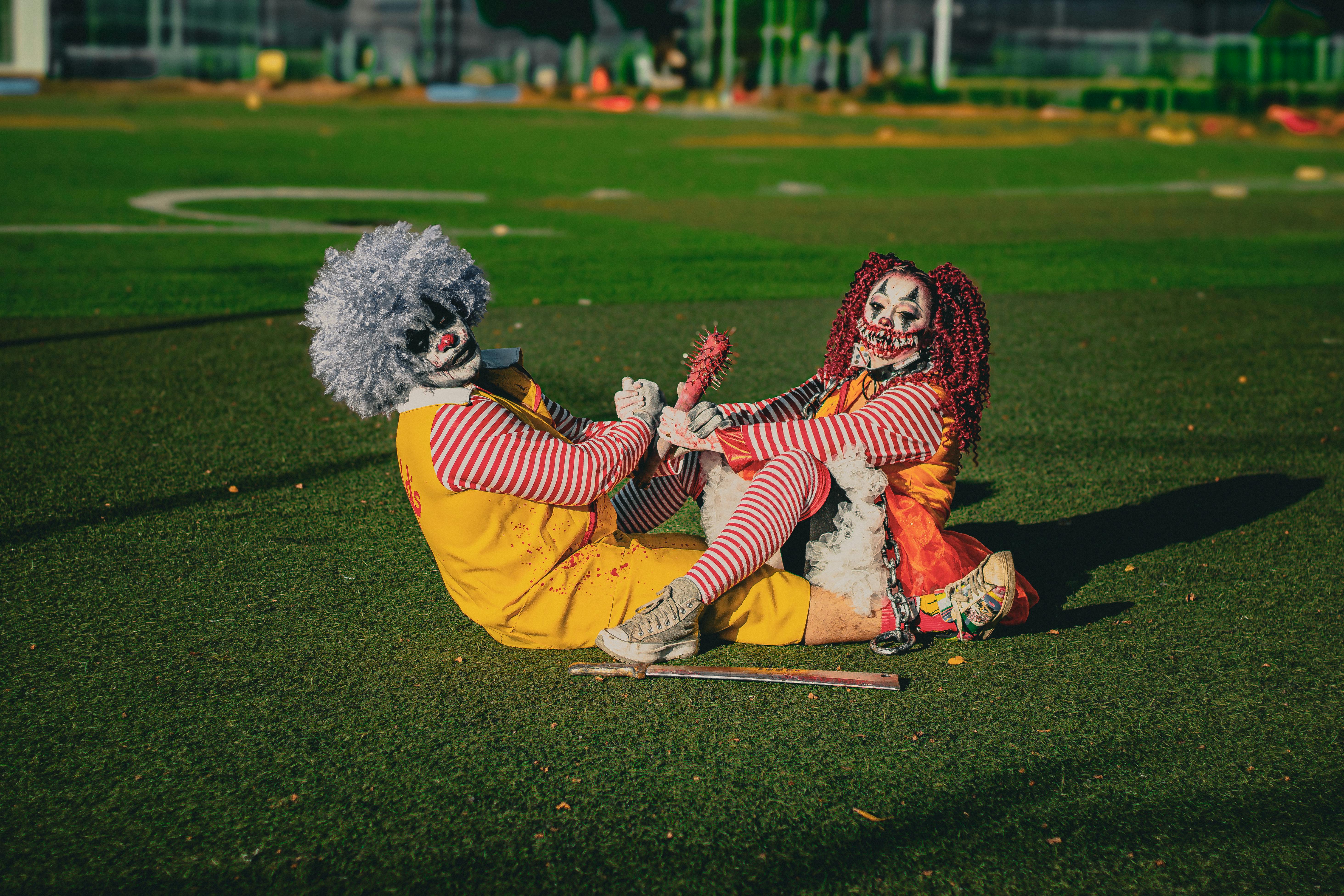 Ücretsiz Güneşli bir alanda şakacı bir şekilde oturan, canlı kostümler giymiş iki korkutucu palyaço. Stok Fotoğraflar