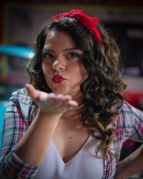 A woman with curly hair and red bow blows a kiss while posing indoors. A playful gesture captured in soft focus.