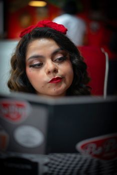 A young woman with vintage makeup and a red headband reading a menu in a retro-styled cafe.