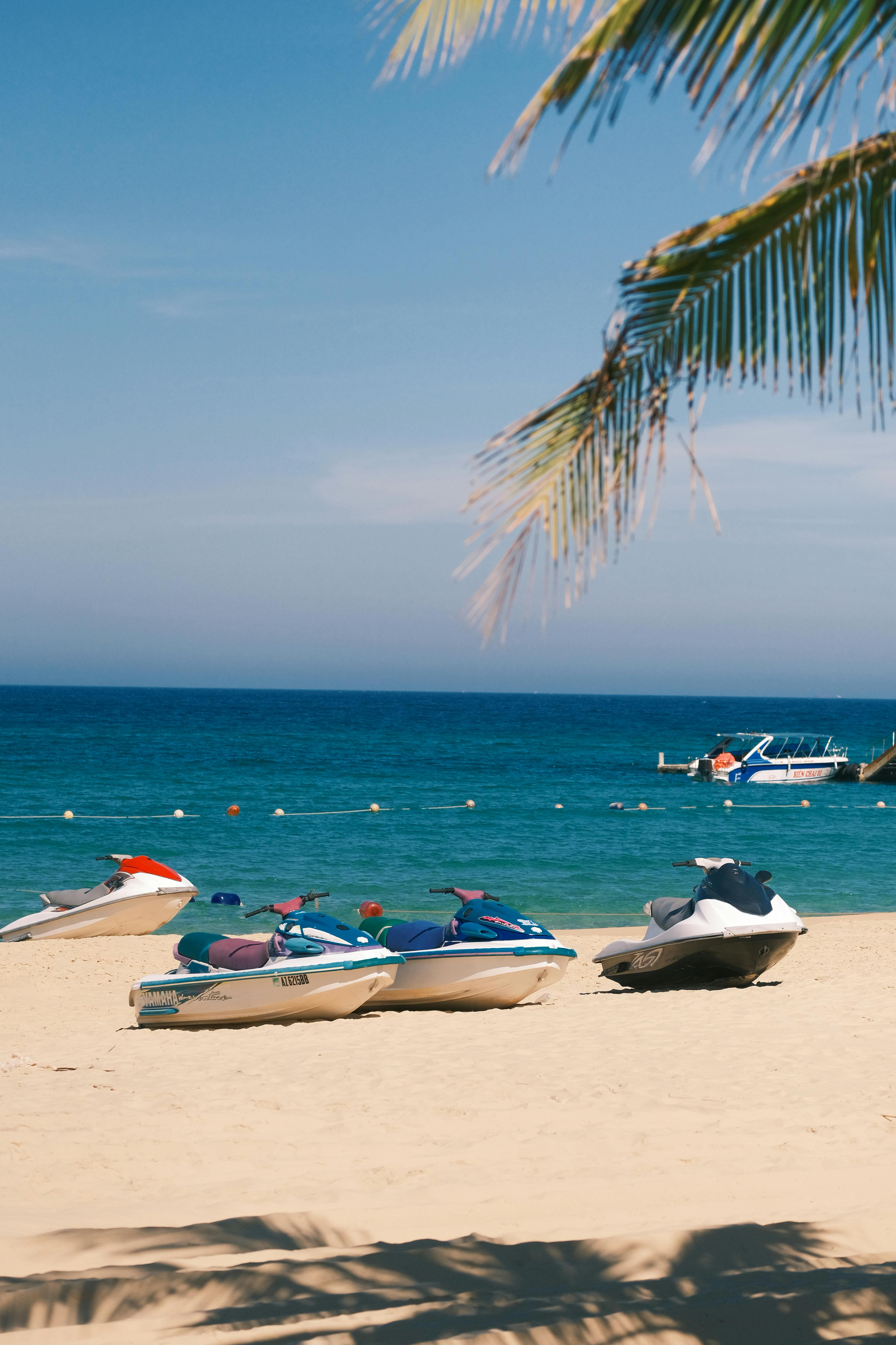 A picturesque sunny beach scene with jet skis on the sand and palm tree shadows.