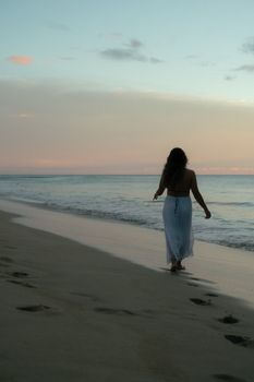 Serene view of woman walking on beach at sunset, leaving footprints in the sand.