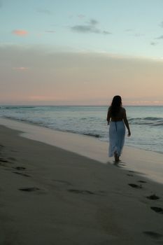 A woman in a white dress walks by the ocean at sunset, leaving footprints in the sand.