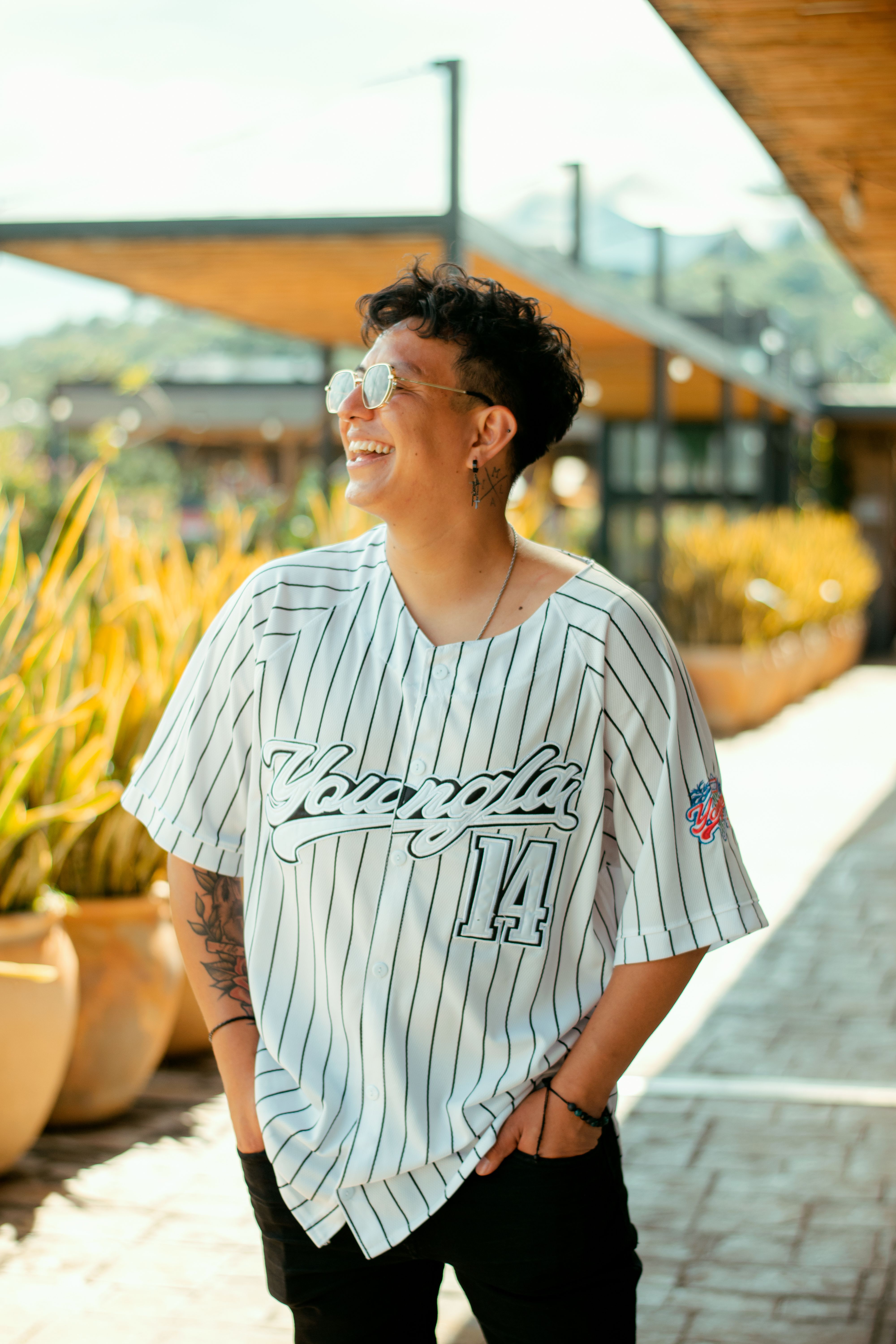 Person with sunglasses smiles confidently outdoors wearing a baseball jersey.
