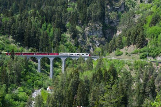 Red train crossing the famous Landwasser Viaduct in Switzerland's picturesque mountain landscape, surrounded by lush greenery.