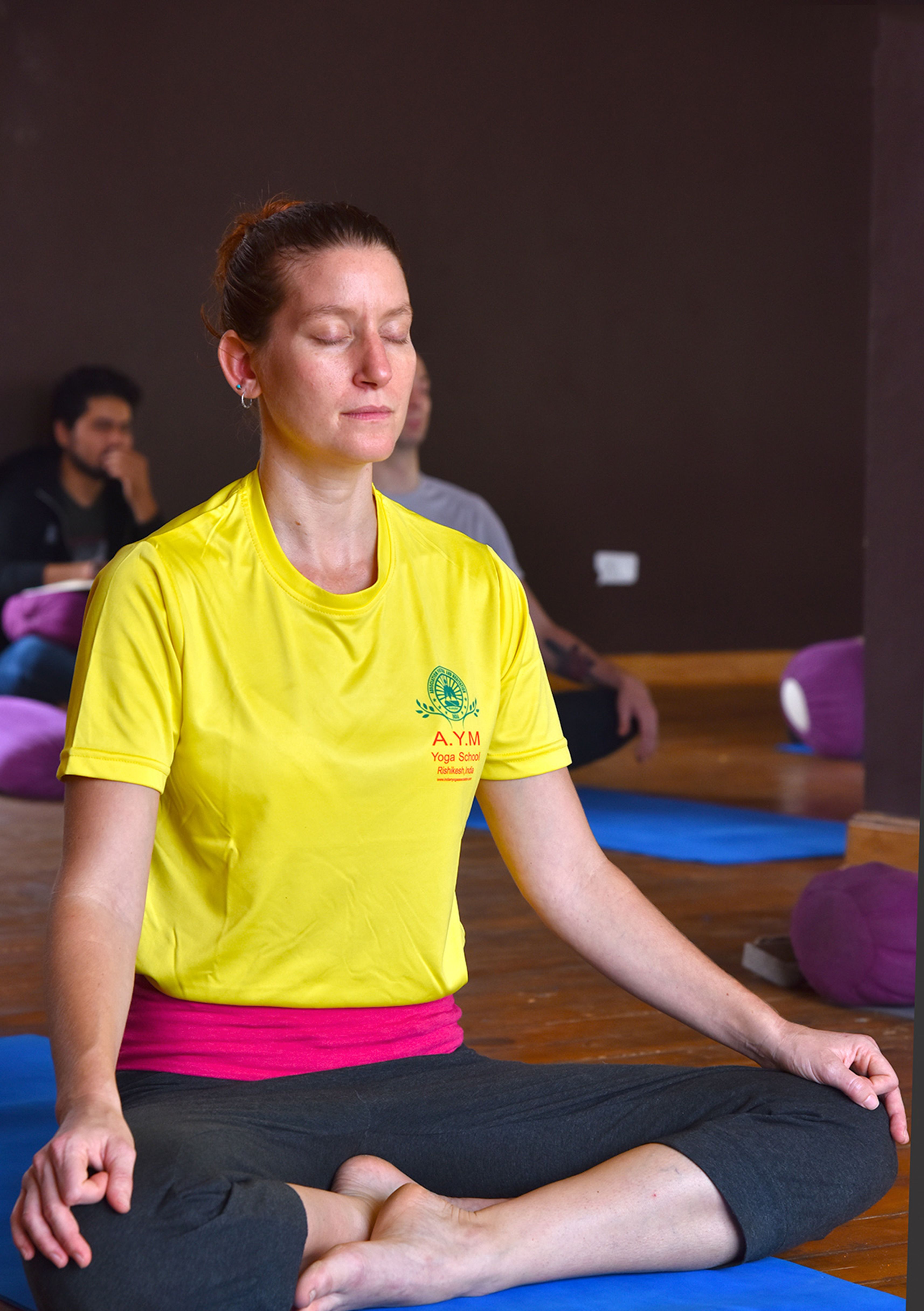 Woman in yellow shirt meditating indoors in Rishikesh, India, seated on a yoga mat.