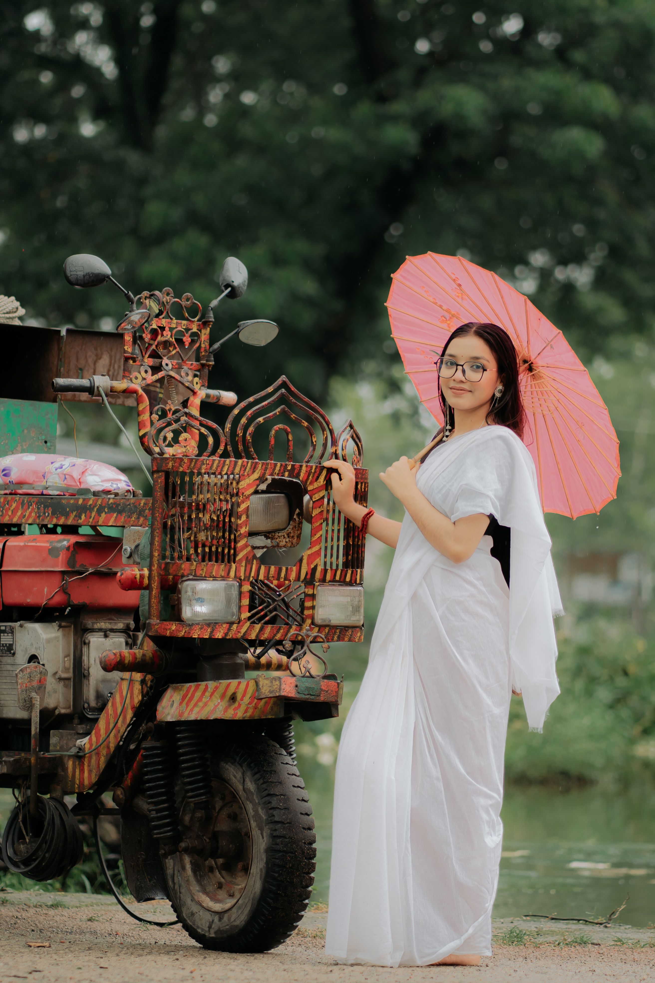 Woman in traditional attire standing by a decorated vehicle with a pink umbrella outdoors.