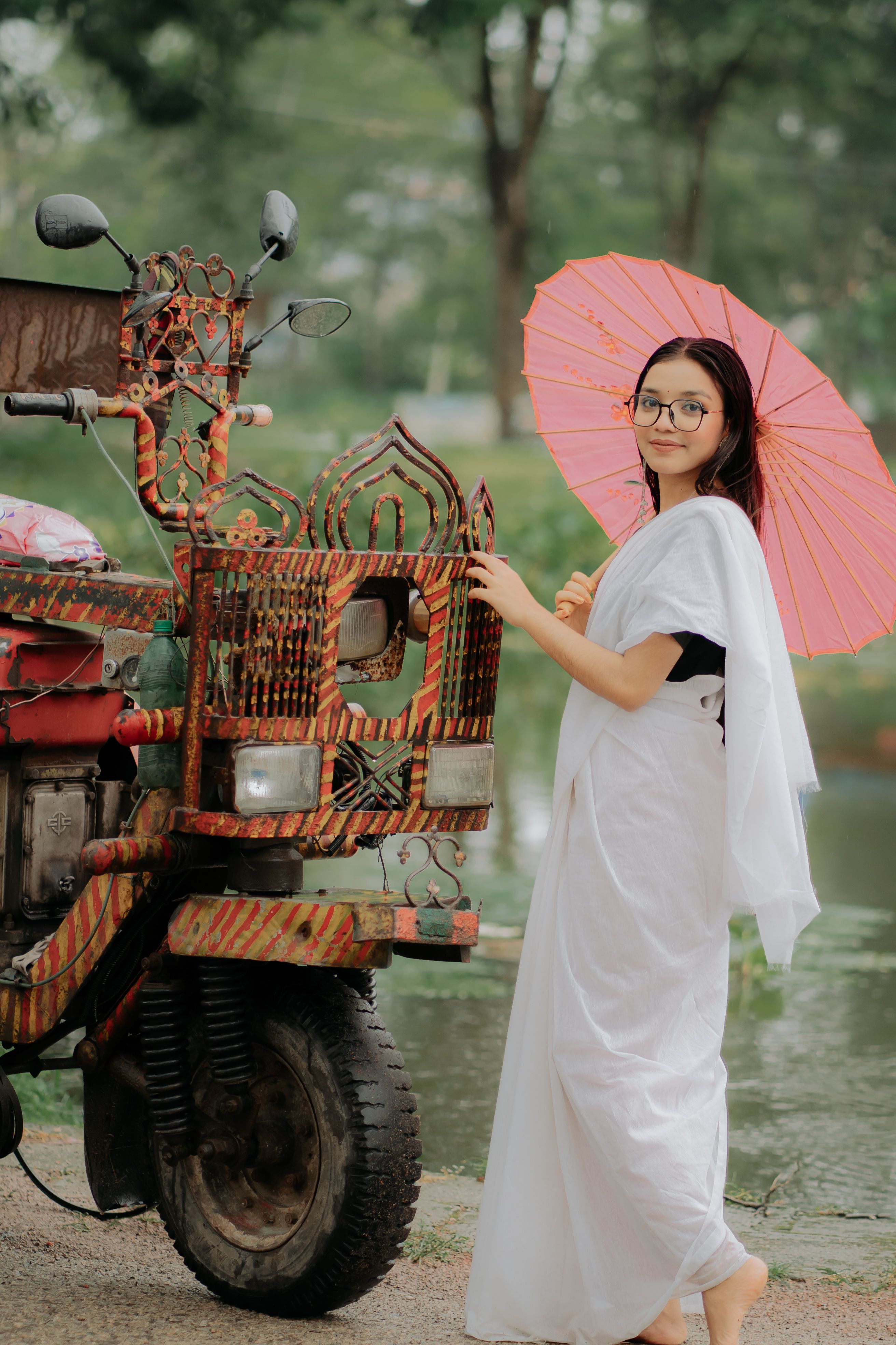 Woman in a white saree holding a pink umbrella next to a decorated vehicle.