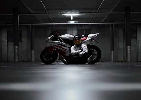Motorcyclist with helmet sits in dimly lit underground parking garage beside high-performance motorcycle.