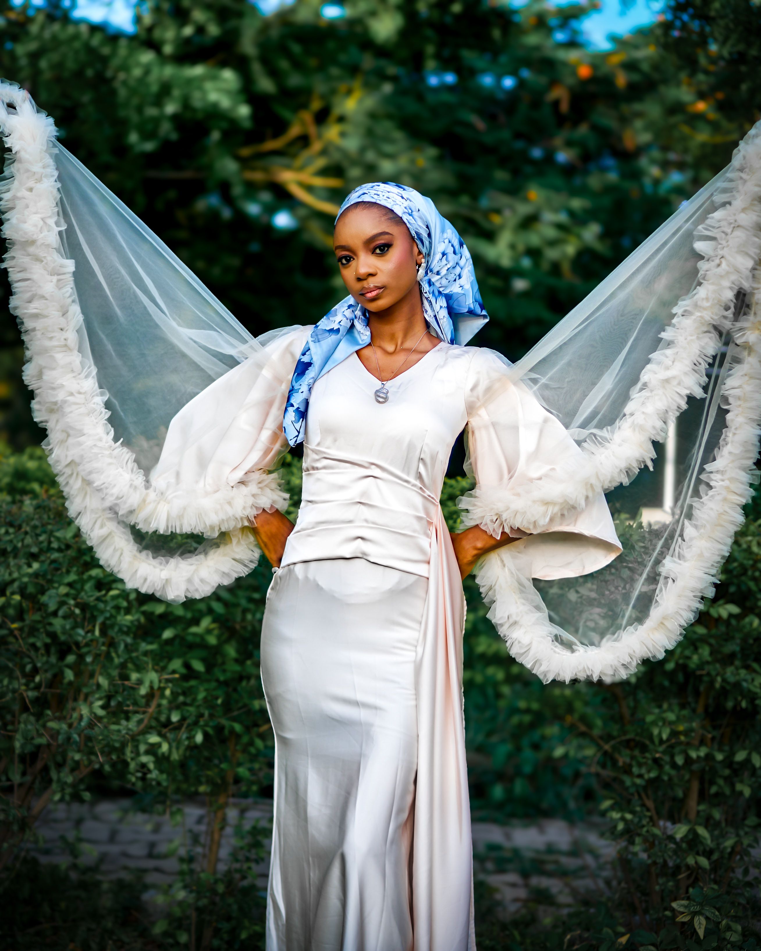 Stylish woman posing in a flowing white dress with feathered sleeves outdoors, exuding elegance.