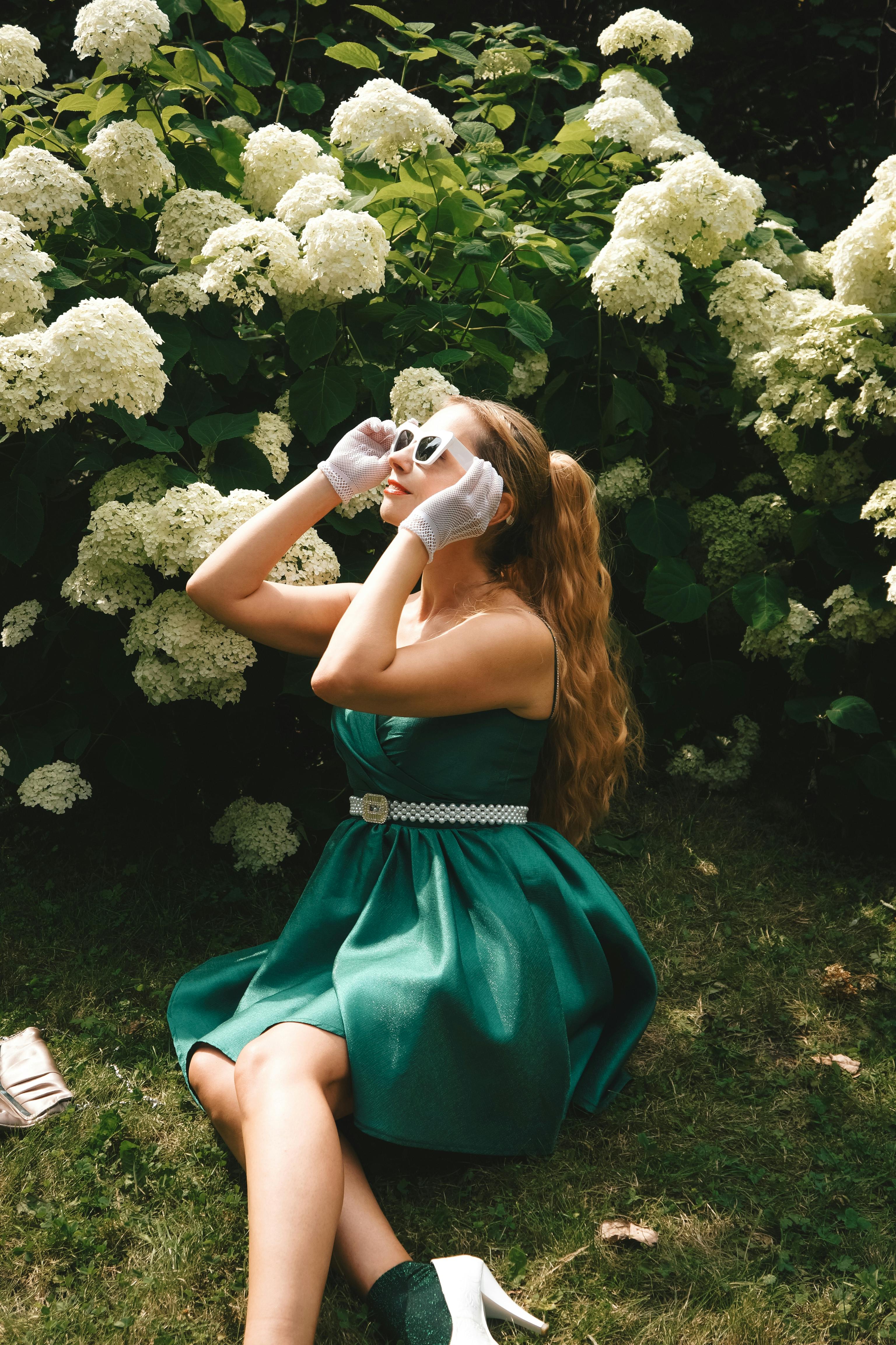 Stylish woman in a green dress and white gloves sitting near hydrangeas in a sunny garden.