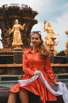 Fashionable woman posing in front of a decorative fountain with golden statues on a sunny day.