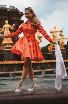 Stylish woman in red polka dot dress posing by ornate fountain outdoors.