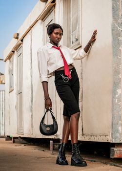Fashionable woman in red tie and boots poses confidently against rustic building. Urban outdoor setting.