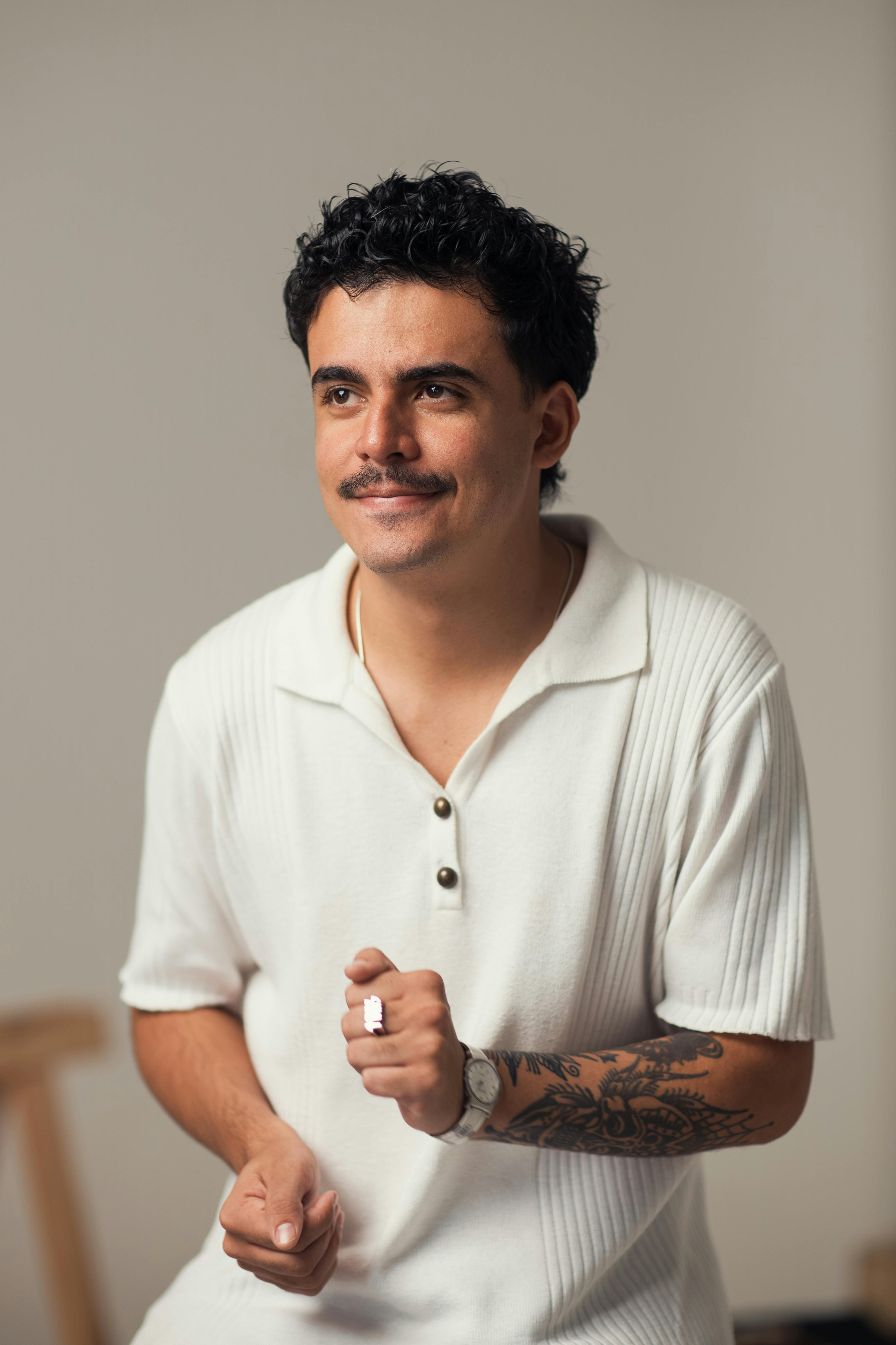 Casual portrait of a young man with tattoos, smiling in a white polo shirt against a simple background.