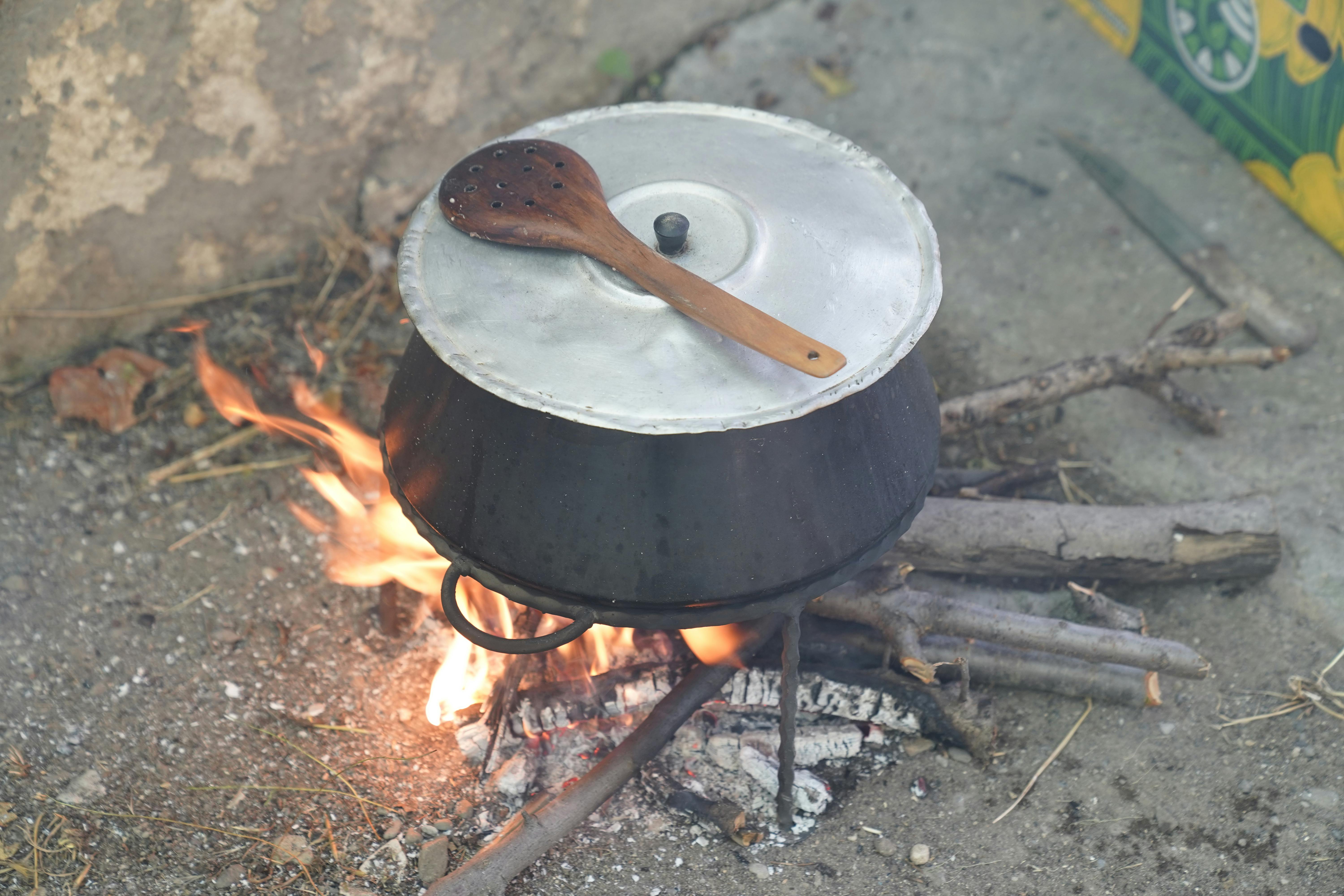 A metal pot with lid sits on an open fire outdoors, with a wooden spoon on top.