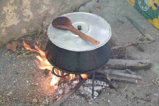 A metal pot with lid sits on an open fire outdoors, with a wooden spoon on top.