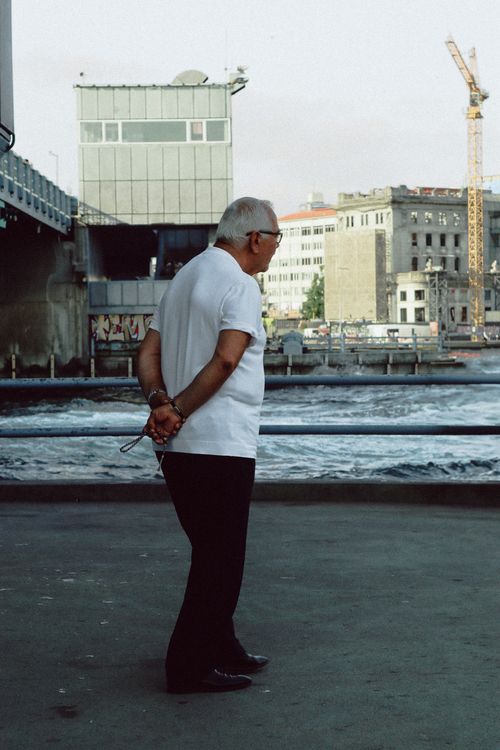 Free A senior man stands contemplatively by an urban river, showcasing city life. Stock Photo
