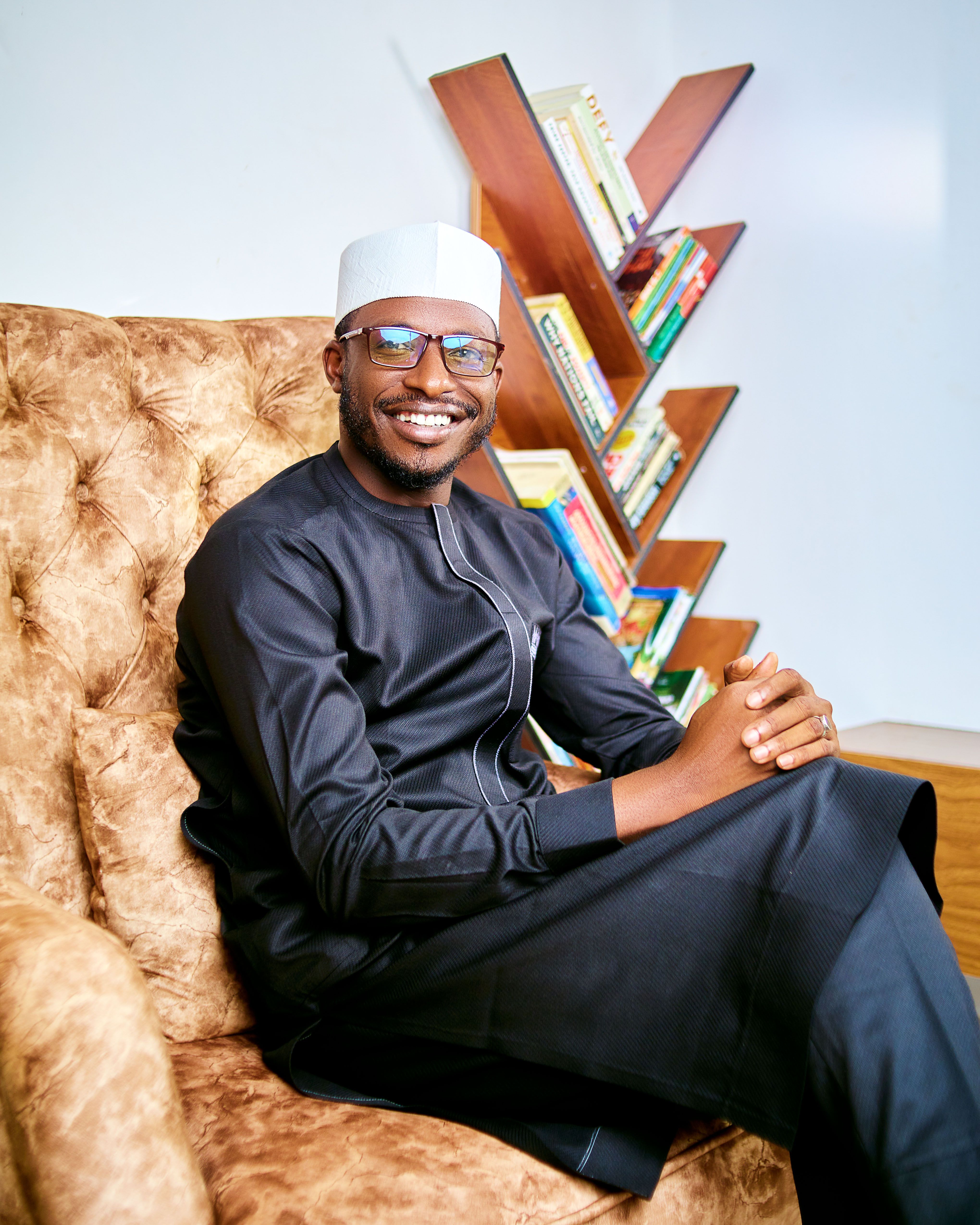 Smiling man in traditional attire seated on a chair beside a geometric bookshelf.