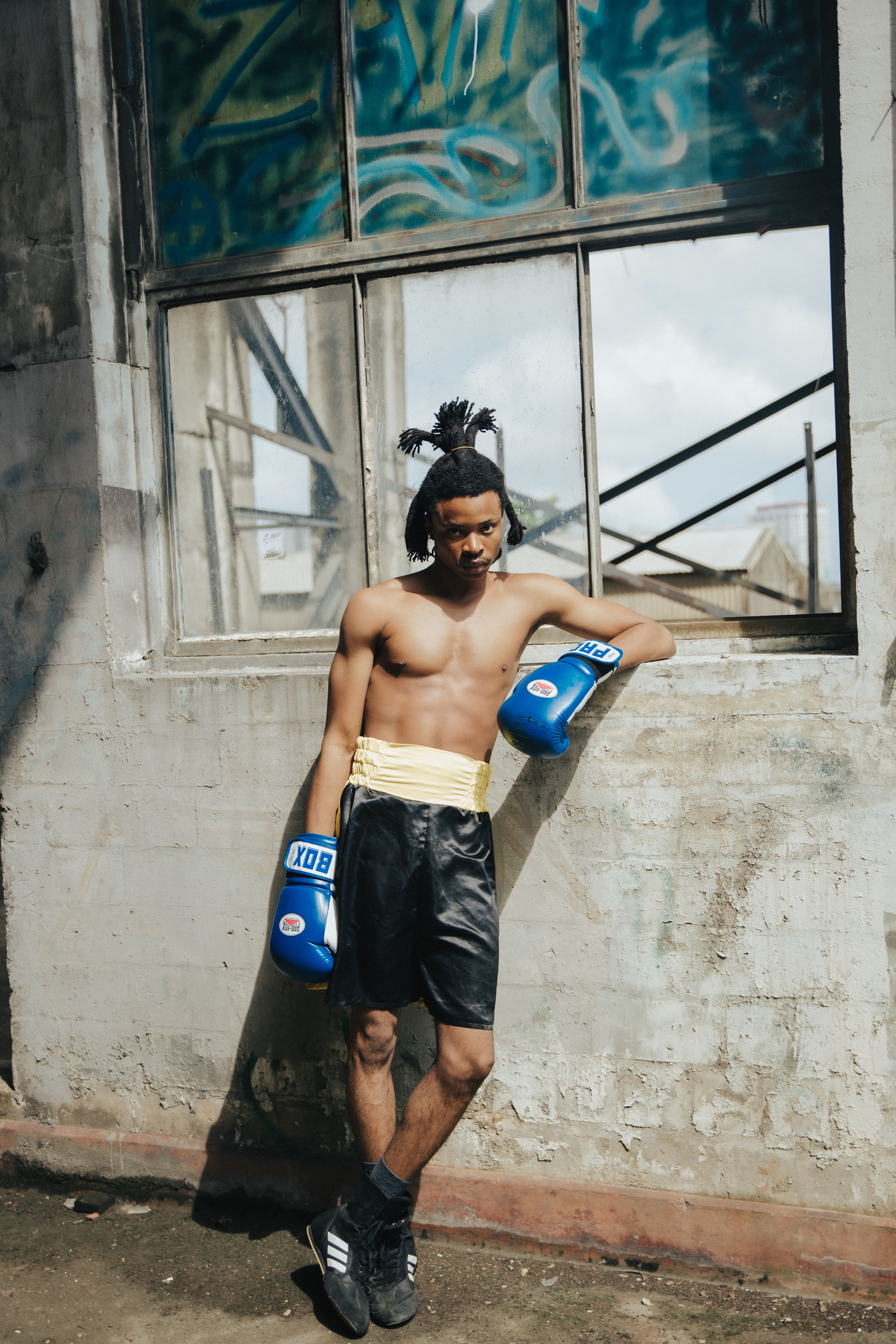 A young male boxer with blue gloves leans casually against a windowed wall outdoors, exuding confidence.