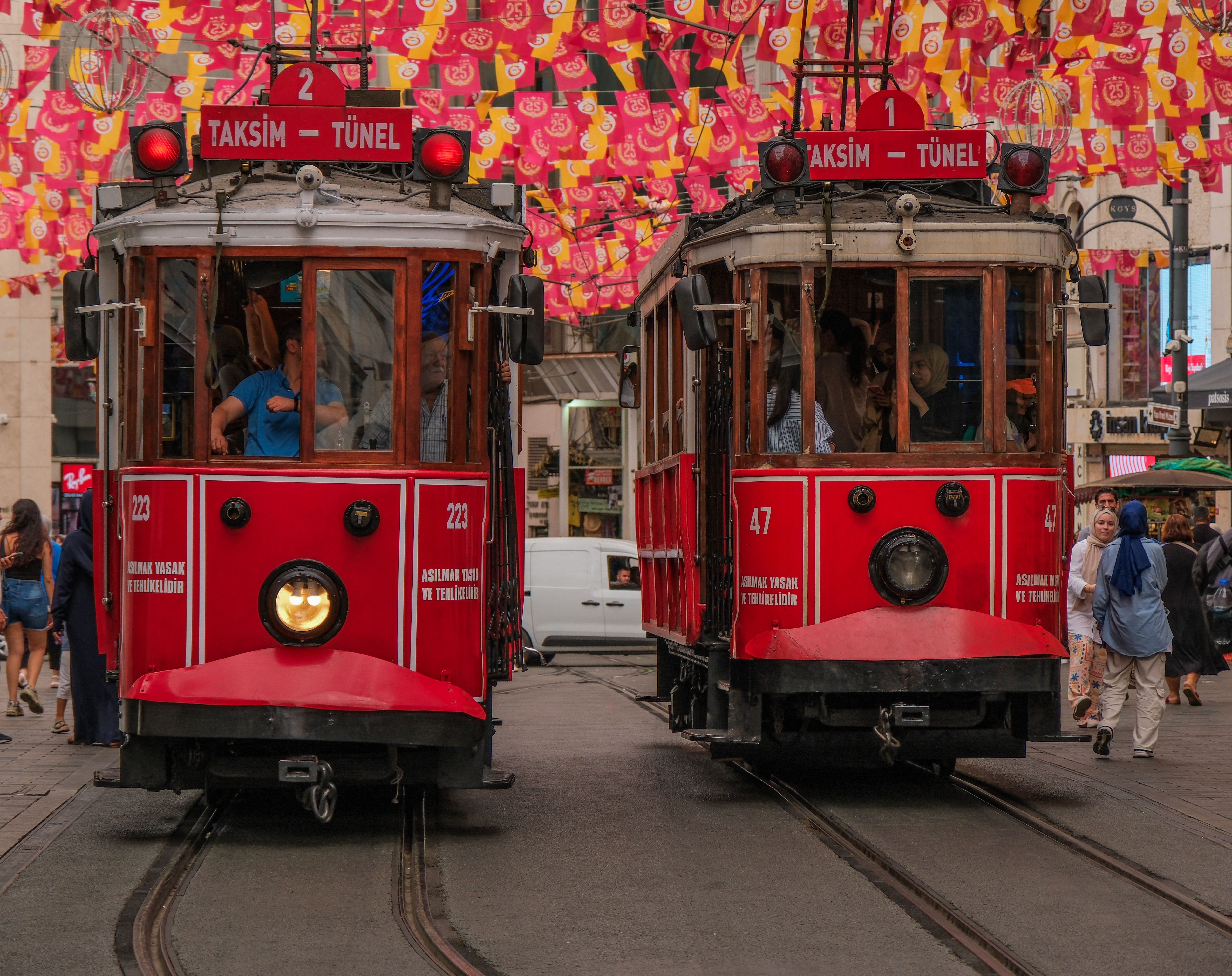 Two red vintage trams signed Taksim–Tünel passing along a street under strings of red flags, with pedestrians beside them