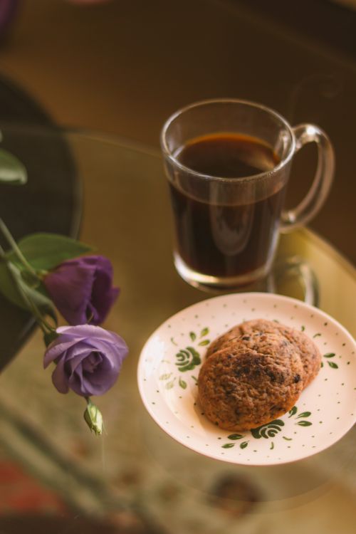 Free Warm and cozy setup featuring a cup of coffee with a cookie and purple flowers on a glass table. Stock Photo
