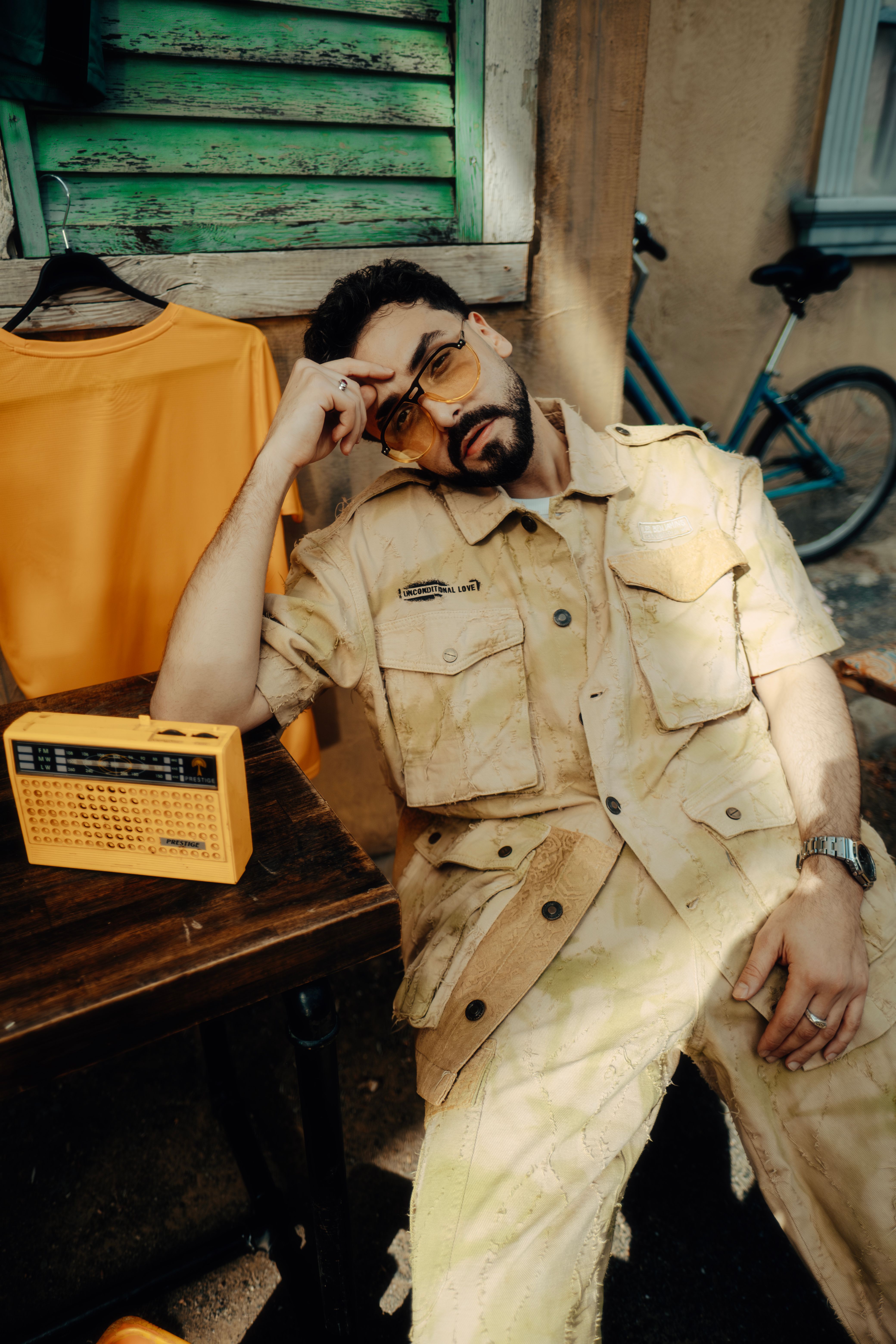 A man in vintage attire relaxes outdoors with a yellow radio and bicycle nearby.
