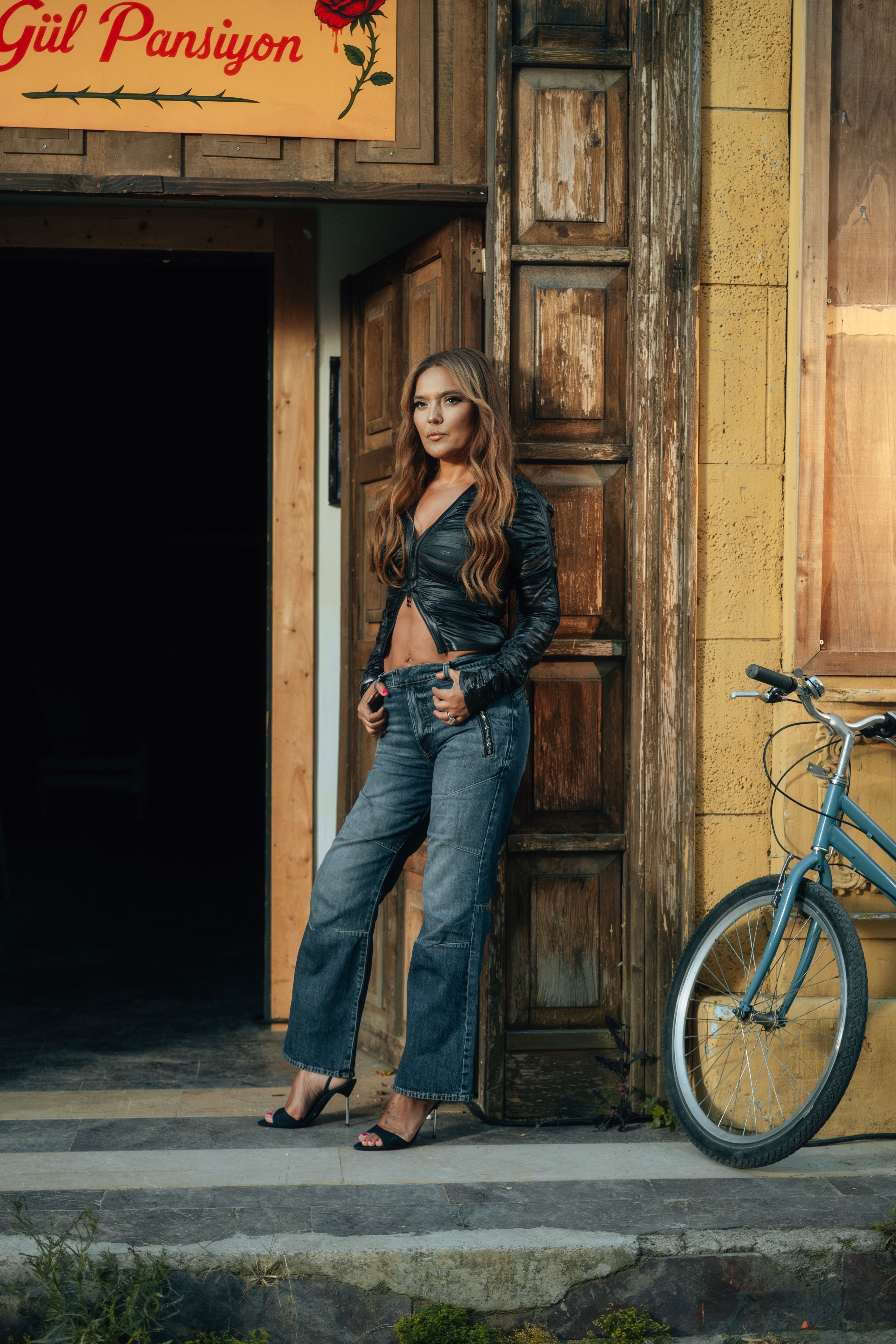 Stylish woman in denim and leather posing outside a rustic building with a bicycle.