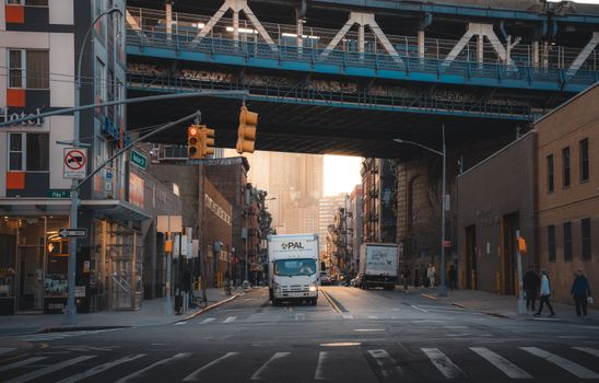A typical street scene in New York City with trucks under a bridge and pedestrians walking.