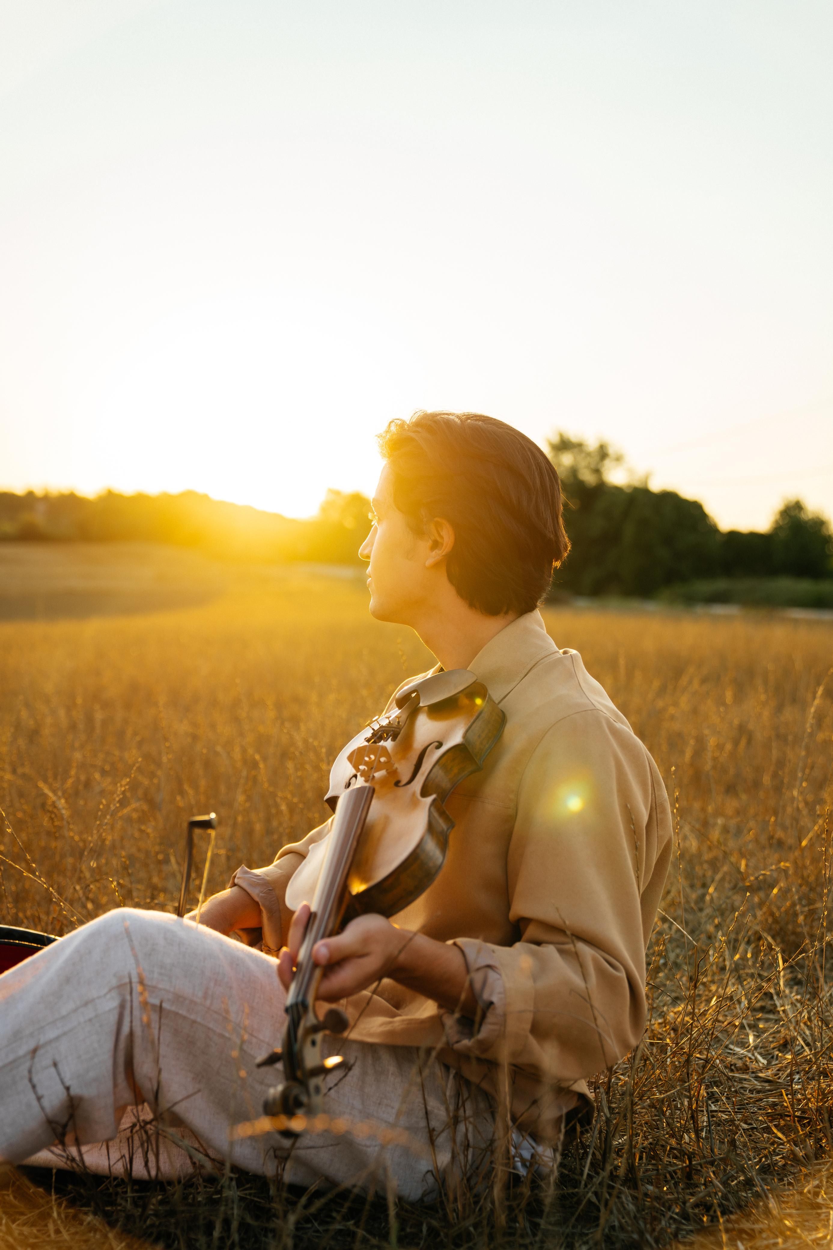 A young man holding a violin, sitting in a field during a beautiful sunset in Istanbul, Turkey.