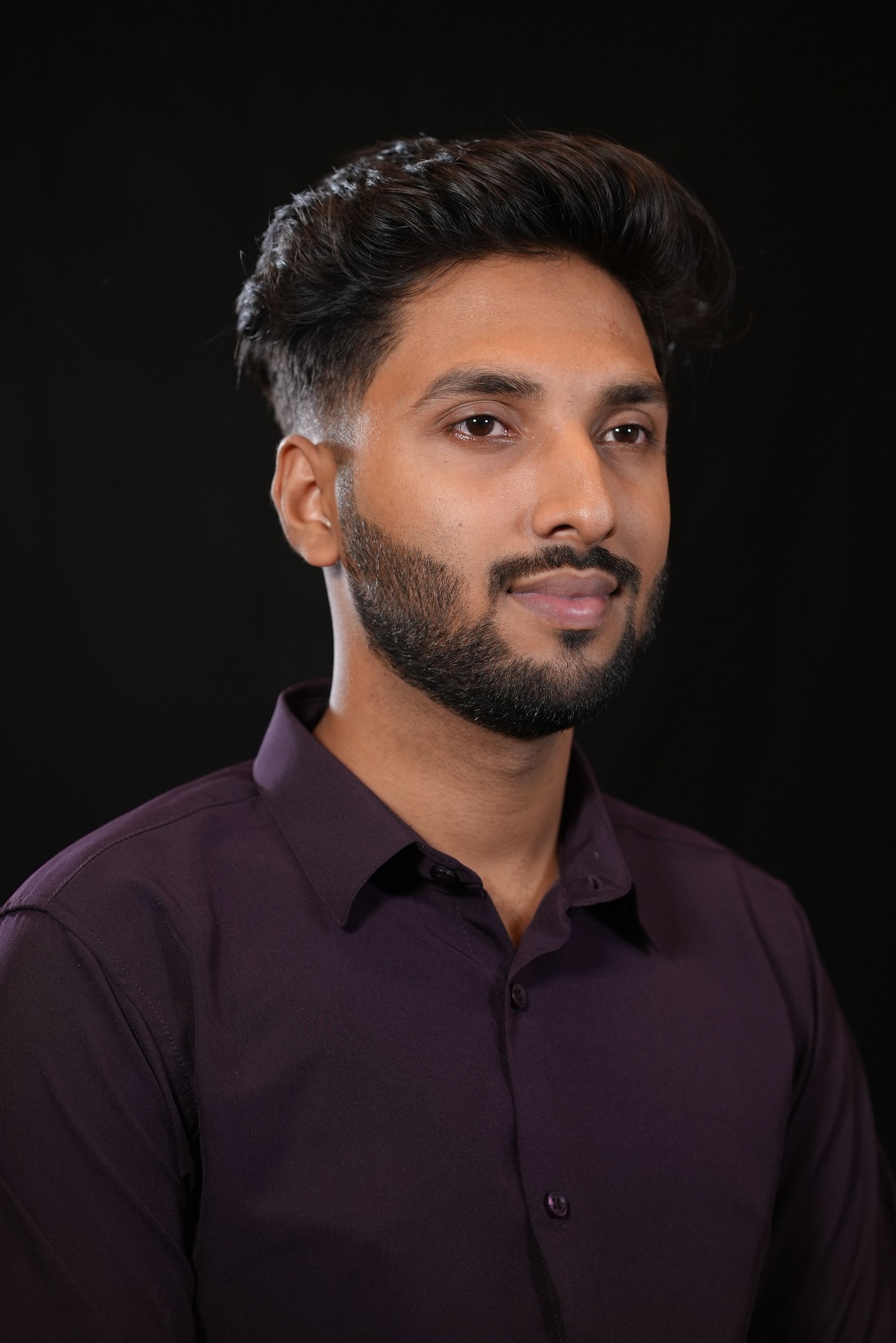 Professional portrait of a stylish South Asian man in formal wear against a dark background.