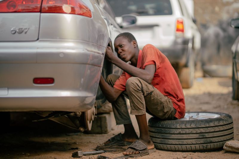 A young mechanic works on a car's underside in an outdoor garage setting.