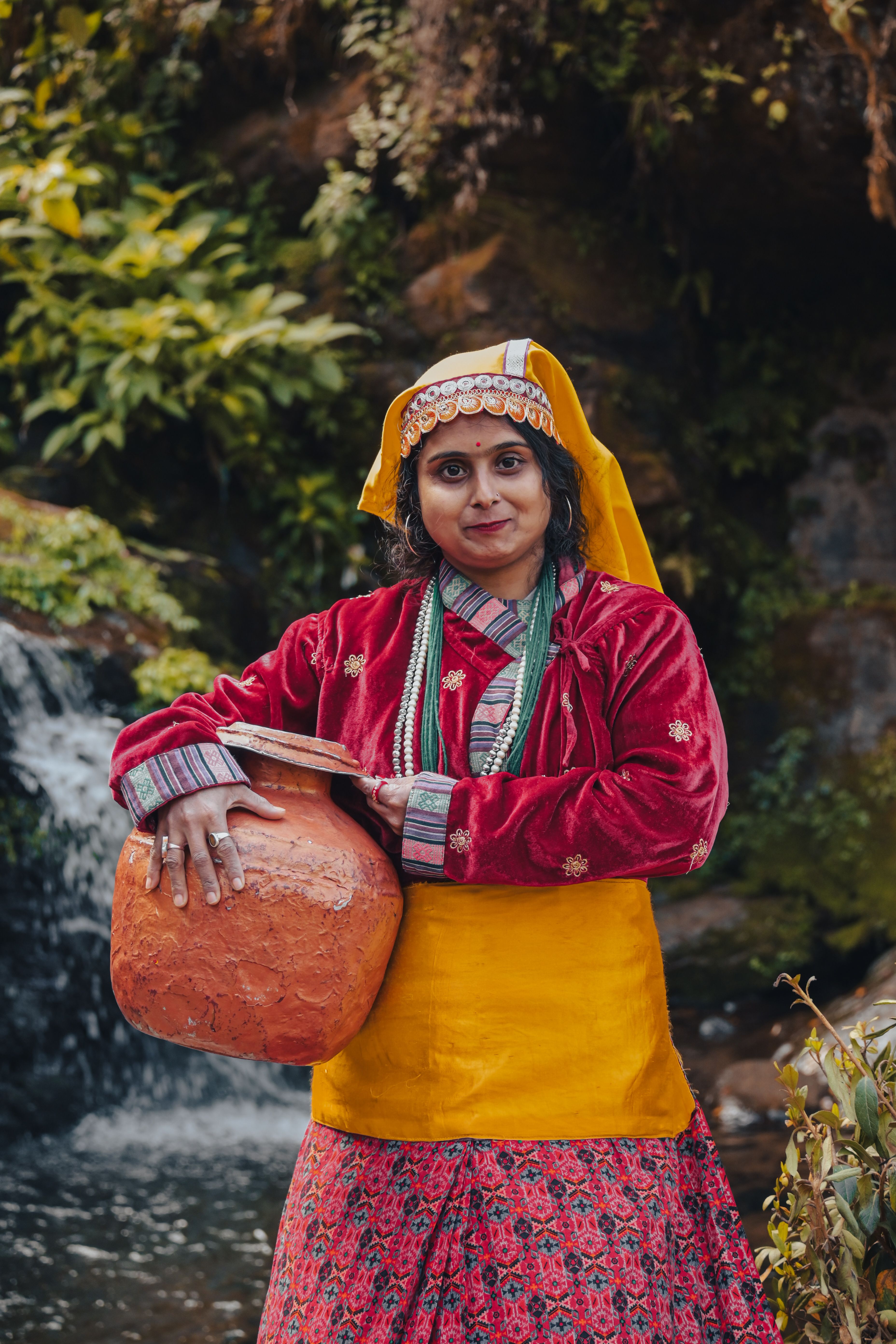 A woman in vibrant traditional Indian clothing holding a clay pot near a waterfall in Darjeeling.