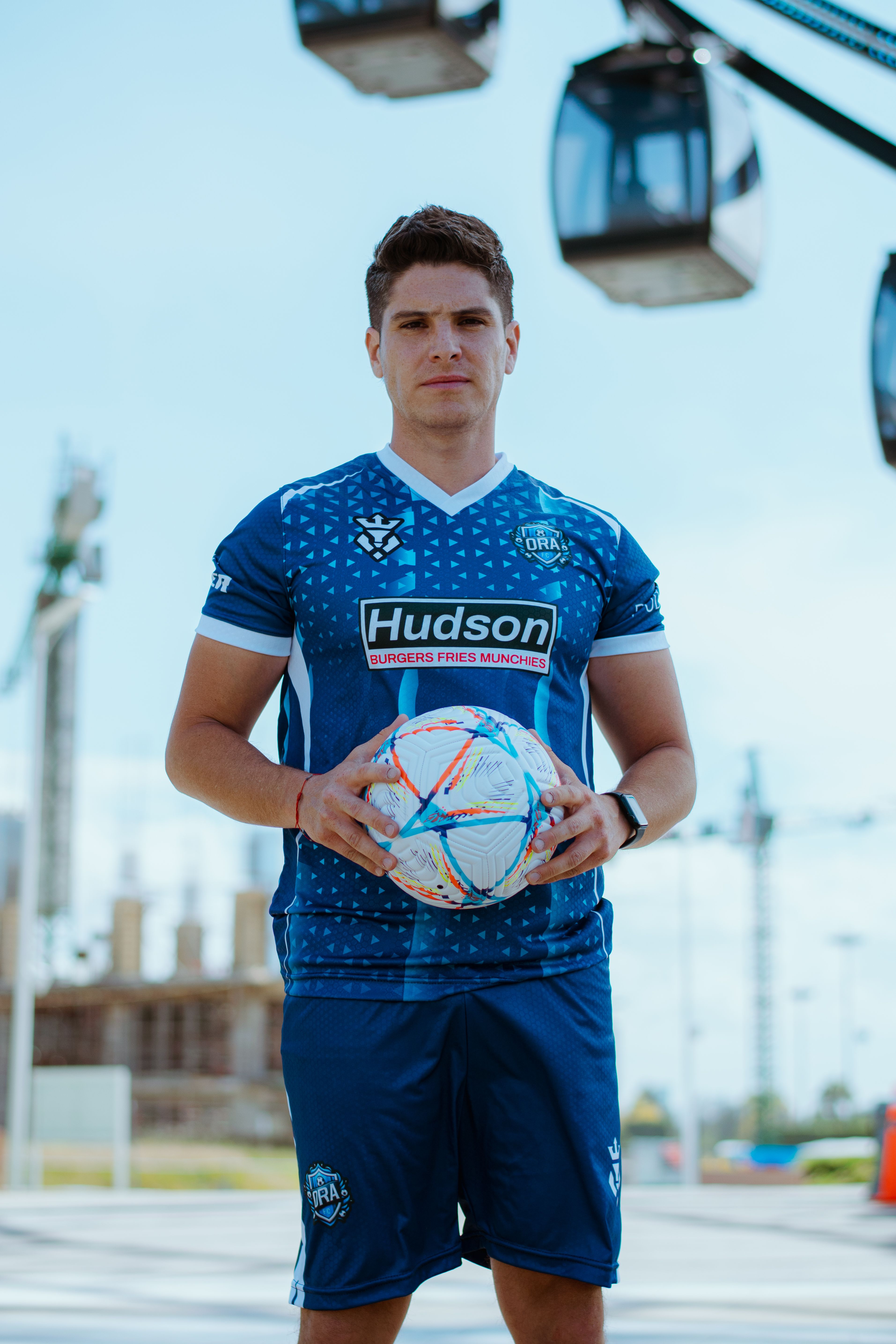 Athletic young man in blue soccer uniform posing confidently outdoors with soccer ball.