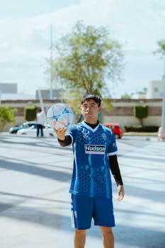 A young male athlete in sportswear holding a colorful soccer ball outdoors in daylight.