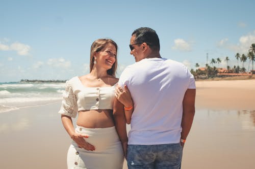 Joyful pregnant couple in trendy outfits embracing and looking at each other with tenderness while standing together on sandy wet seashore in tropical resort
