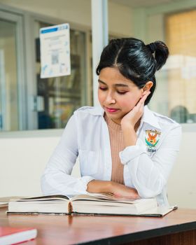 A young Hispanic woman in a university setting, focused on reading a textbook inside a study area.