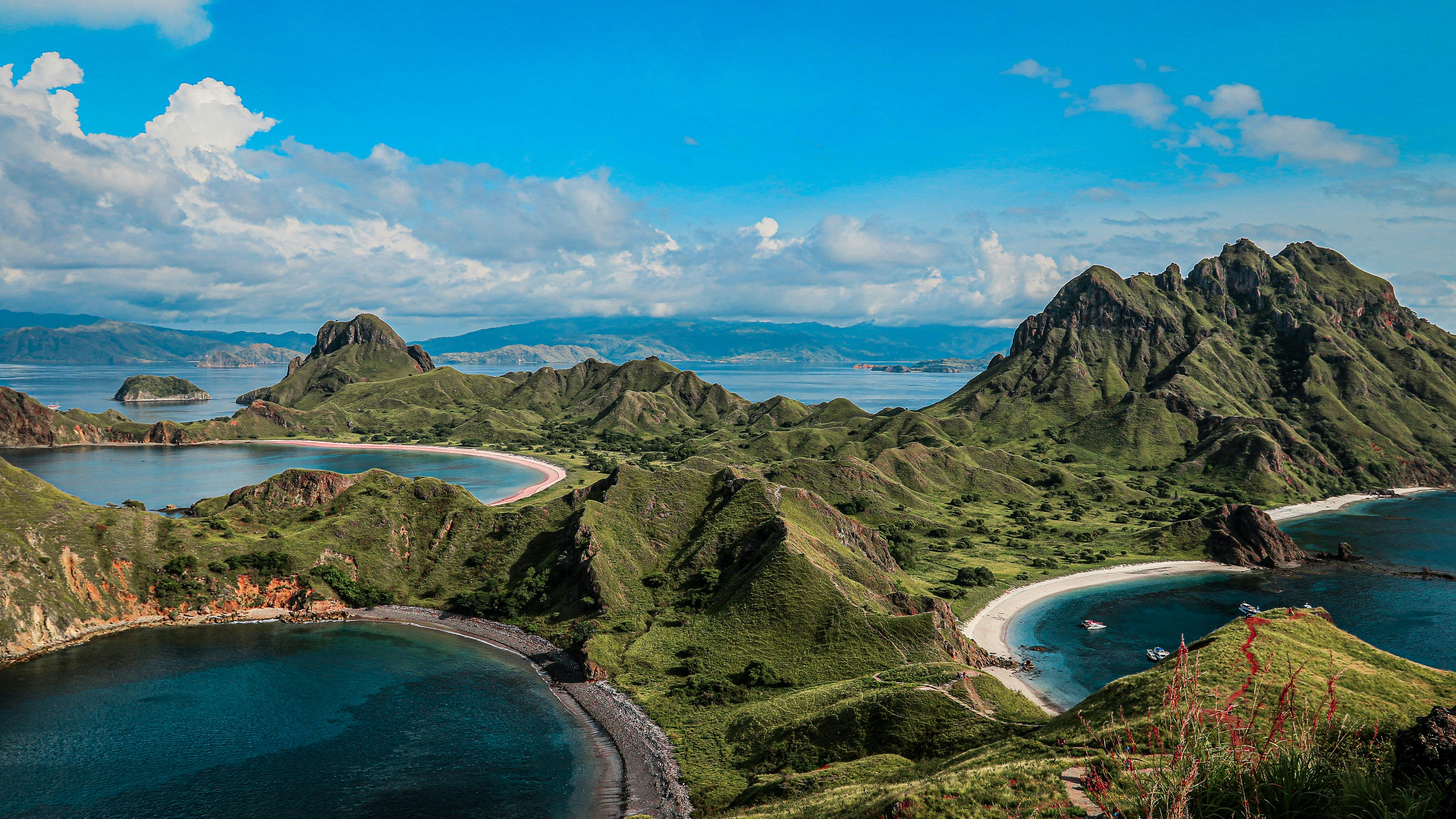 Meanwhile, a panoramic view of Labuan Bajo showcases lush green hills and rugged peaks above twin crescent beaches and turquoise bays, with small boats anchored near the shoreline under a bright blue sky.