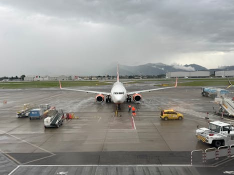 A commercial airplane on a wet tarmac at an airport during rainy weather with mountains in the background.
