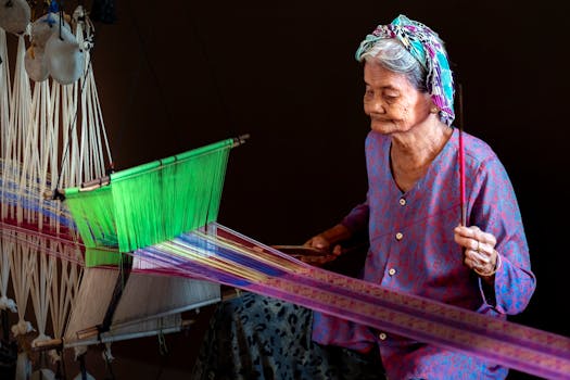 Elderly woman skillfully weaving colorful textile on a loom inside a dim room.