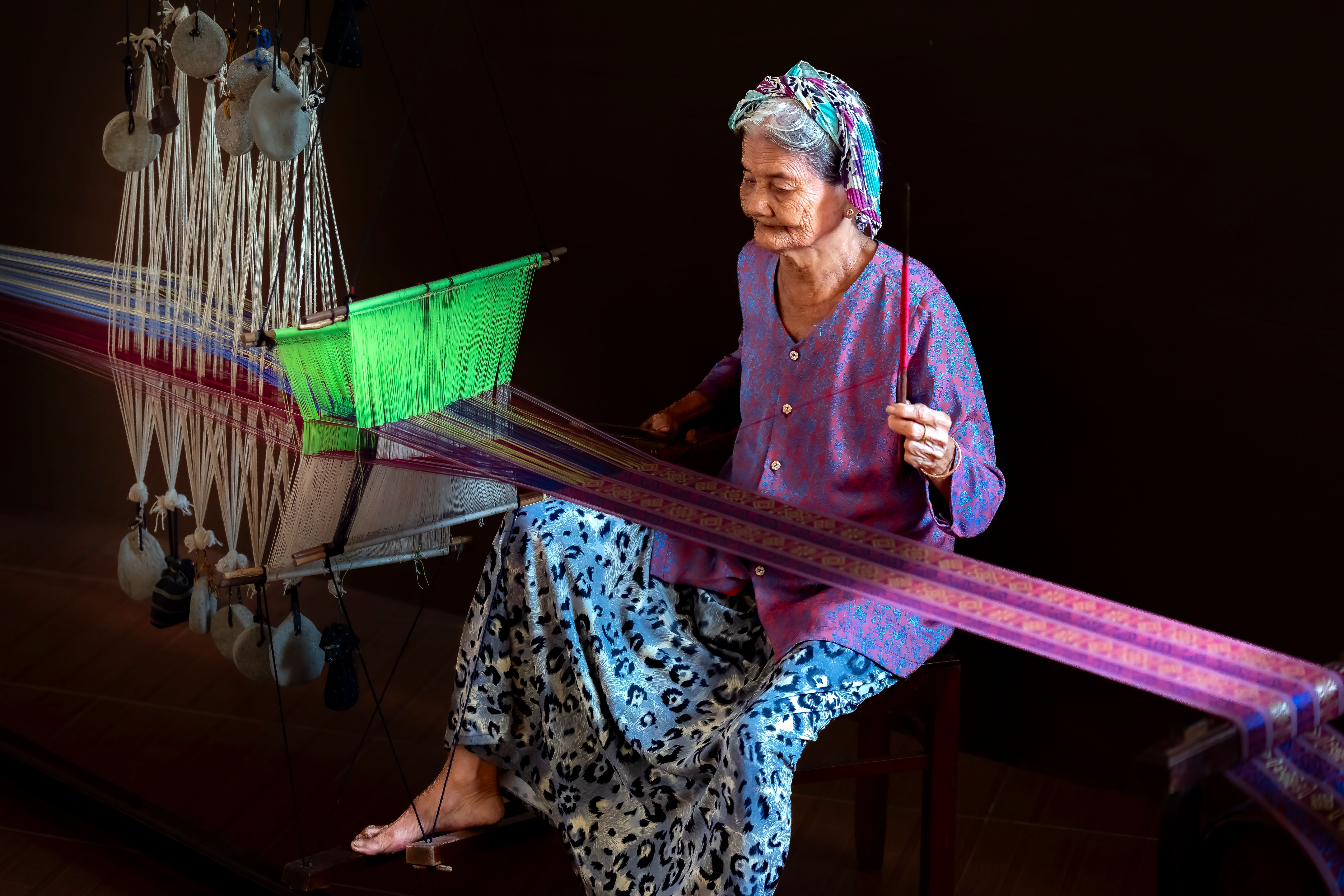 Elderly woman skillfully weaving vibrant fabric on a traditional loom indoors.