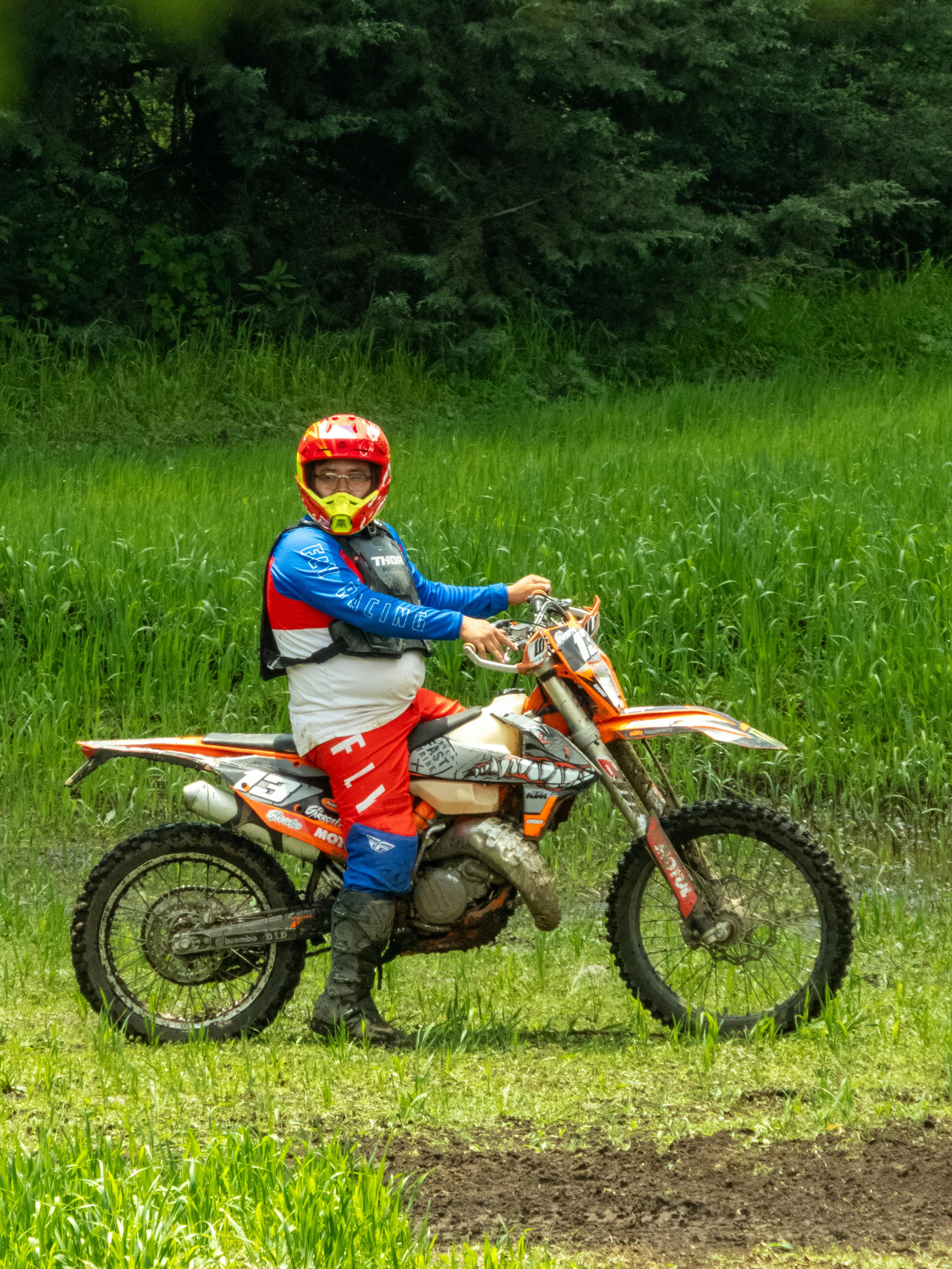An off-road motorcyclist in full gear pauses on a dirt trail surrounded by greenery.