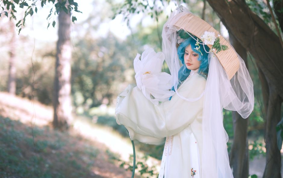 Cosplayer in elaborate costume with blue hair and floral accents set in a serene outdoor environment.
