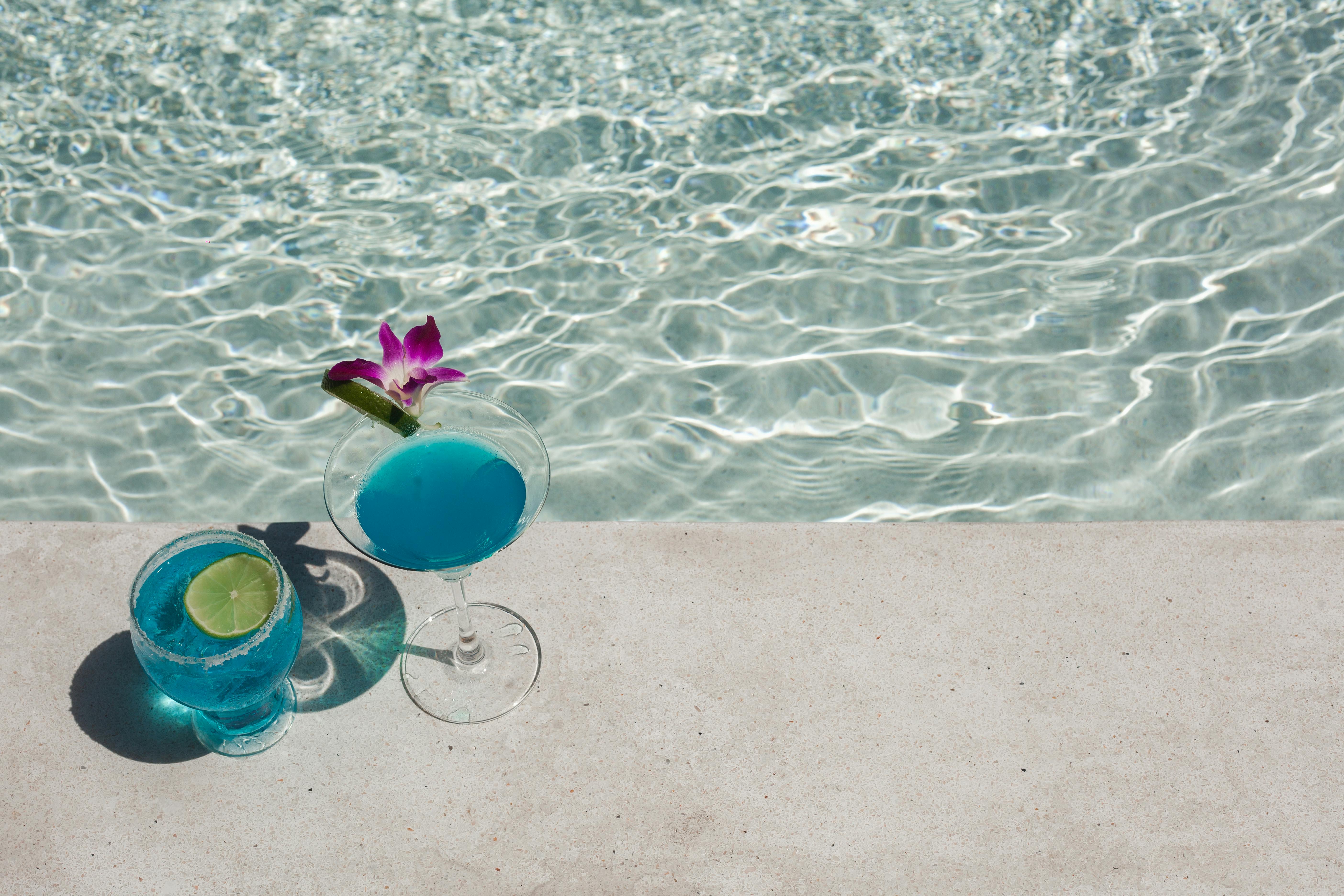 Two blue cocktails with lime and orchid by a sunlit poolside, perfect for a tropical vacation.